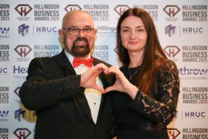 Hillingdon Business Awards 2025. A man and woman are standing in front of the photo wall. Each is holding up one hand to make the shape of half a heart.