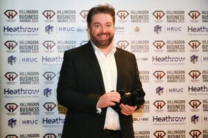 Hillingdon Business Awards 2025. Matt Wareham standing in front of the photo wall. He's holding a camera as he was the videographer.