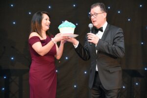 Hillingdon Business Awards 2025. The host handing over a large cupcake to the lady who made it. She owns the business Tany's Bakes.